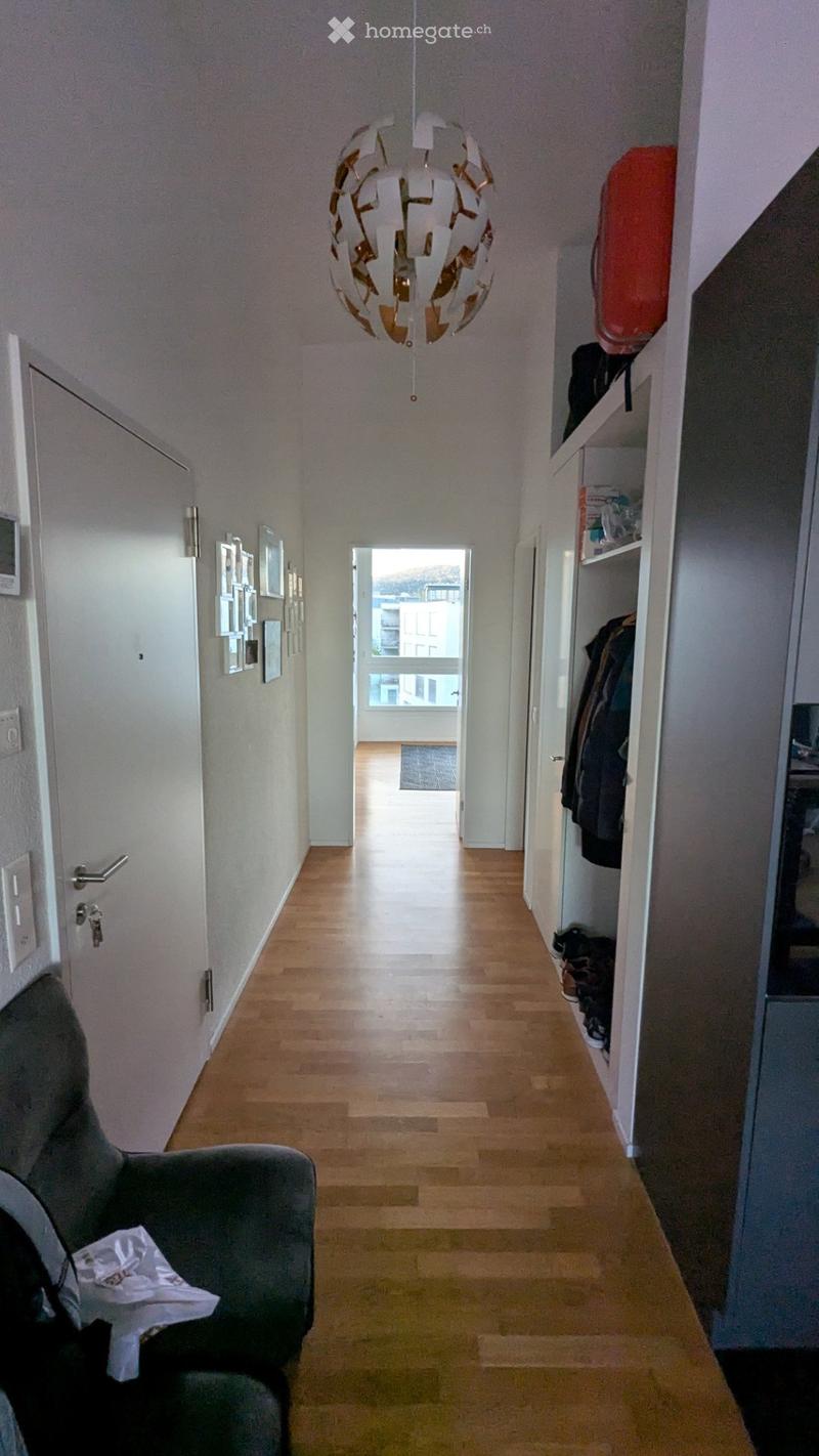 A well-lit hallway with wooden flooring, white walls, and a modern chandelier. A coat rack with jackets and shoes is visible on the right, and a grey armchair with a white cloth is on the left. The hallway leads to an open area with large windows and a vi