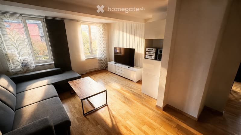 Modern living room with wooden flooring, large windows, grey sectional sofa, wooden coffee table, flat-screen TV on a white stand, and a stereo system with speakers.
