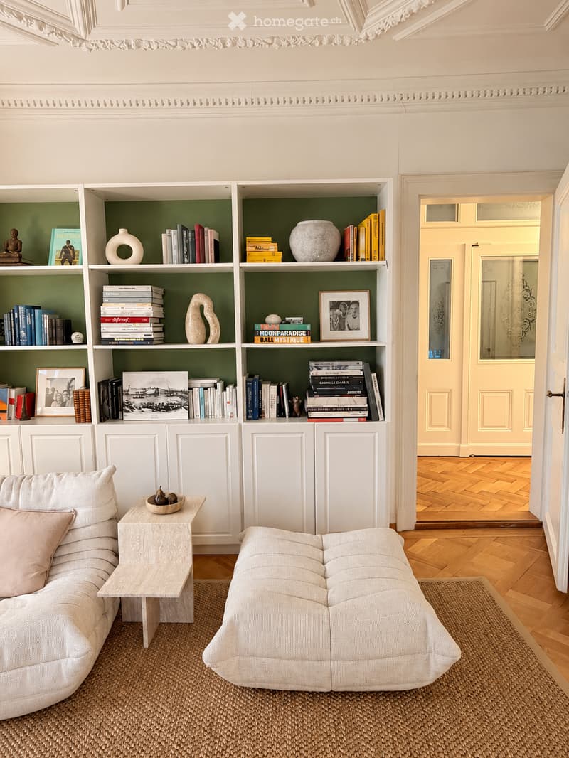 A modern living room with a light-colored sofa, ottoman, side table, and a bookshelf filled with books and decorative items. The room features wooden flooring and a woven rug.