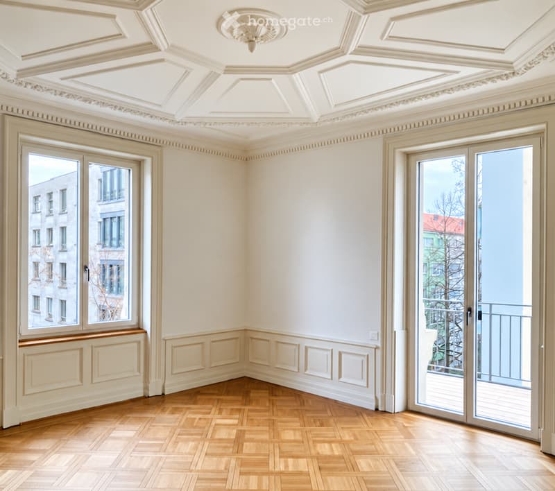 Empty room with wooden parquet flooring, white walls, decorative molding, and large windows with views of neighboring buildings. A sliding glass door leads to a balcony.