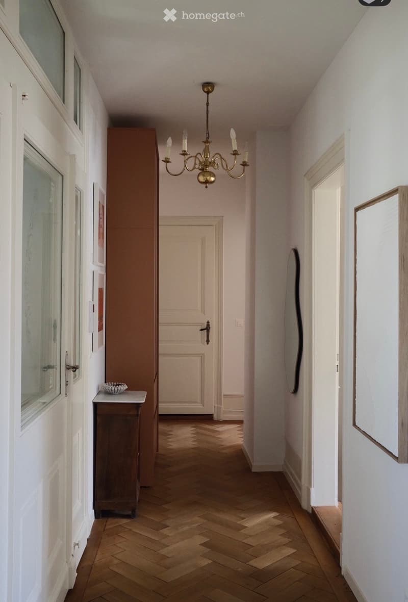 A narrow hallway with wooden herringbone flooring, white walls, and a chandelier hanging from the ceiling. There are several doors and a small console table with a decorative bowl on it.
