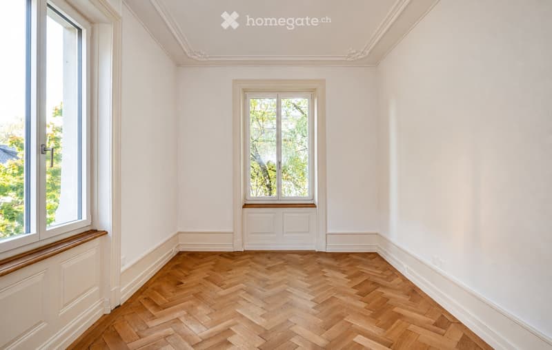 Empty room with white walls, herringbone wooden flooring, and two windows with white frames. The room features decorative crown molding and a small built-in bench beneath one window.