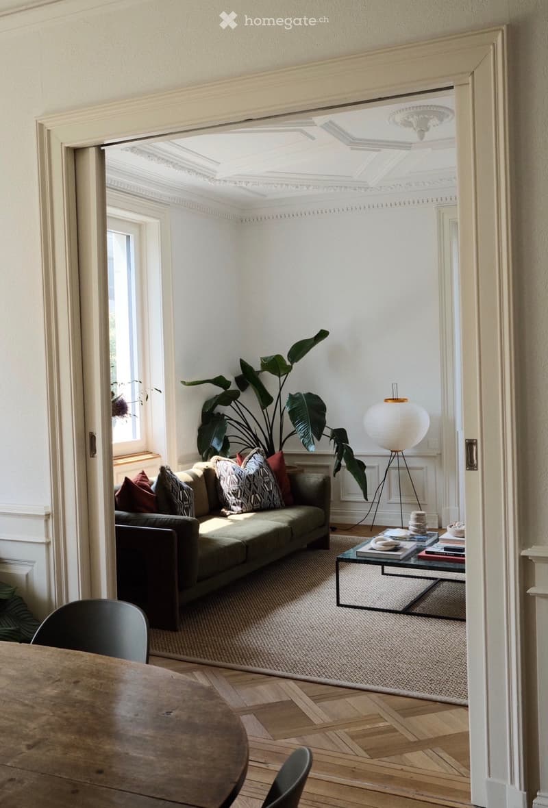 Elegant living room with a green sofa, patterned cushions, a coffee table with books, a floor lamp, and a large leafy plant near the window.