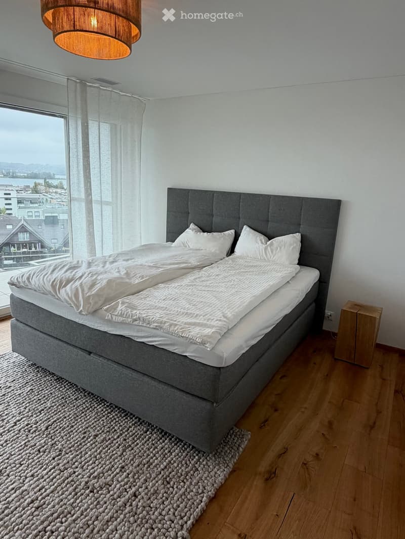 Modern bedroom with a large double bed, grey headboard, white bedding, wooden nightstand, and a textured rug on a light wooden floor. Large window offers a scenic view.