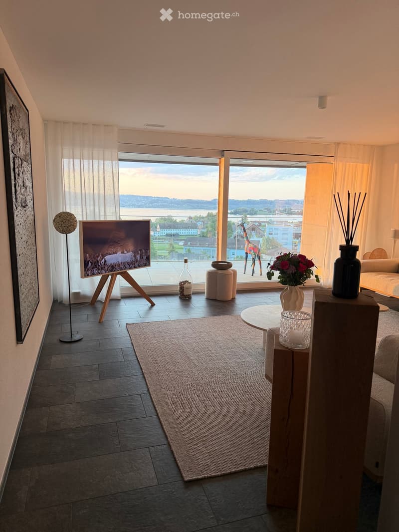 Modern living room with large windows offering a scenic view, featuring a sofa, coffee table, floor lamp, television on a stand, decorative vase, stool, and a round table with a glass centerpiece.