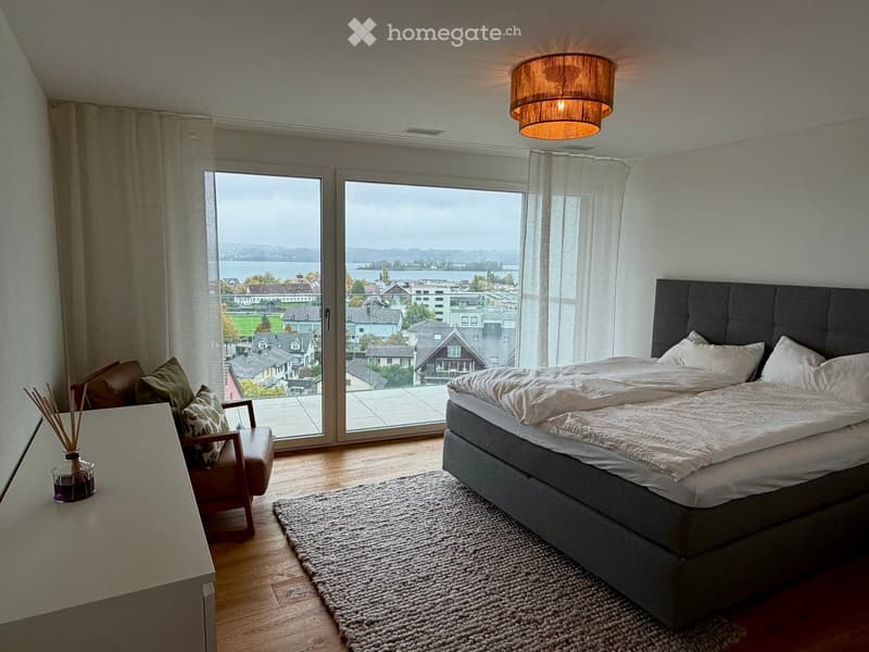 Modern bedroom with large windows offering a scenic view, featuring a bed, a wooden chair, a small table, a rug, and a hanging lamp.