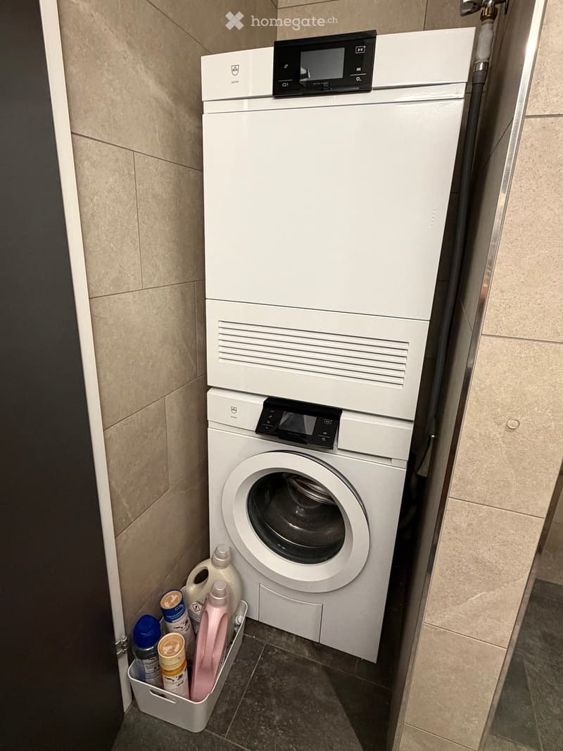 A compact bathroom with a stacked washing machine and dishwasher unit, surrounded by cleaning supplies and a small basket on the floor.