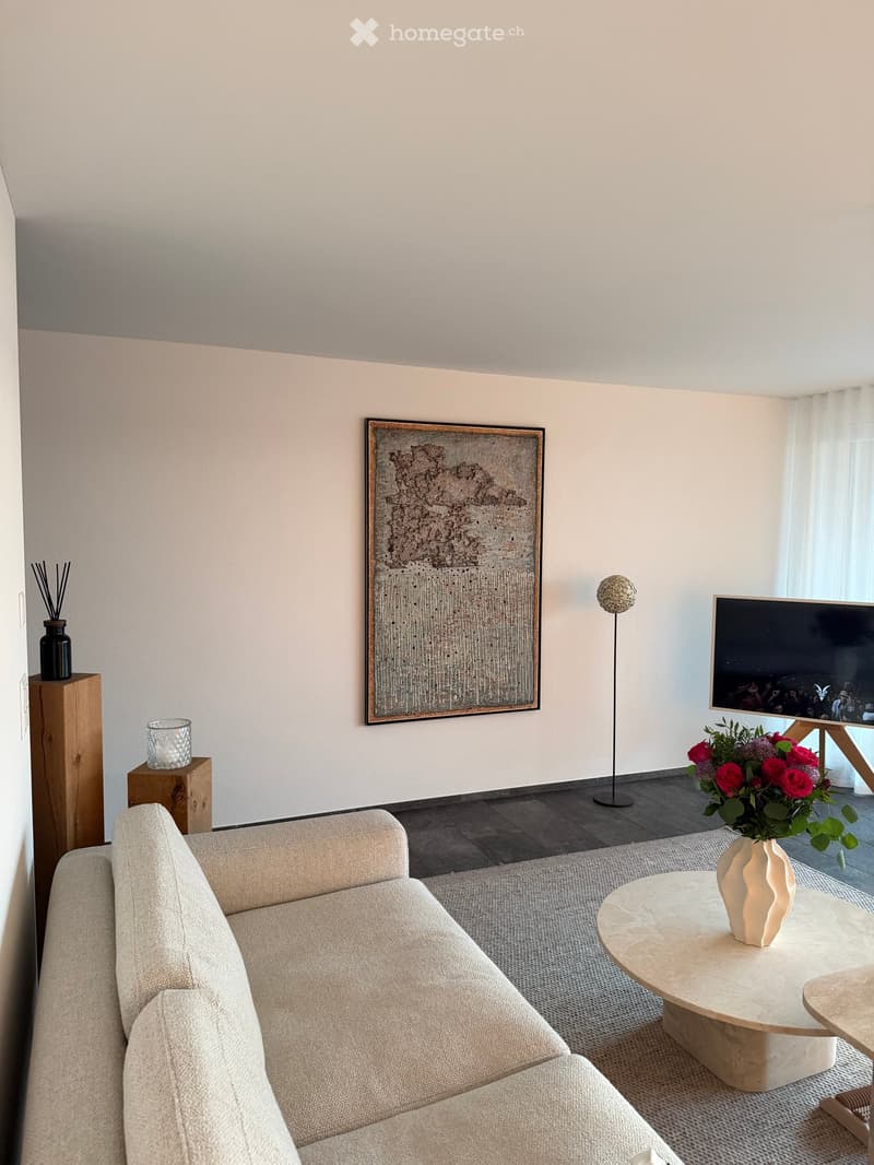 Modern living room with a light-colored sofa, a round coffee table with a decorative vase, a floor lamp, a large screen, and a picture frame on the wall.