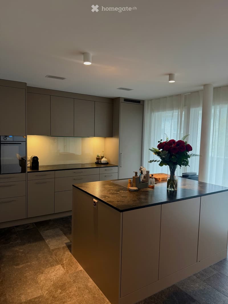 Modern kitchen with grey cabinets, a central island, and a dark countertop. Features include an oven, sink, and various small appliances and decorative items on the counter.