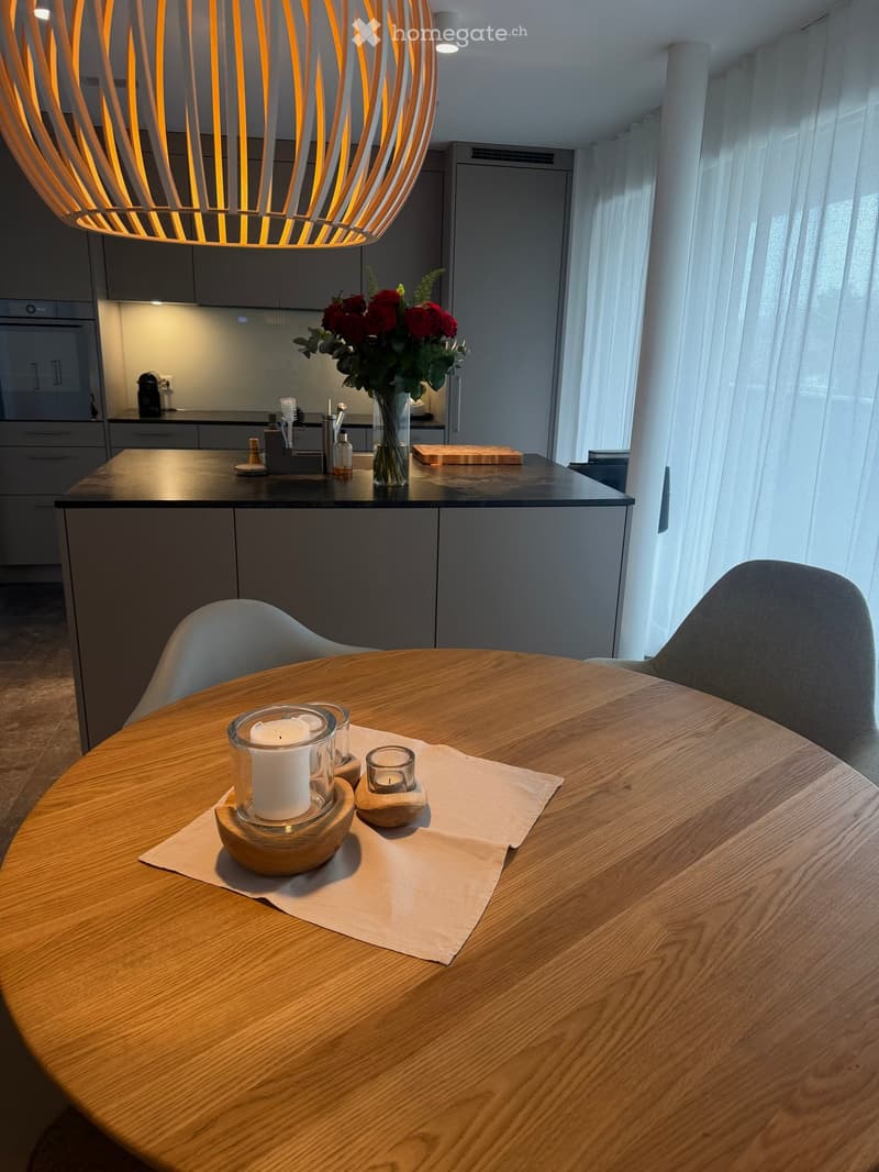 Modern kitchen with a round wooden dining table, two chairs, a vase with red flowers, and a candle on the table. The kitchen features grey cabinets, a black countertop, and a hanging pendant light.