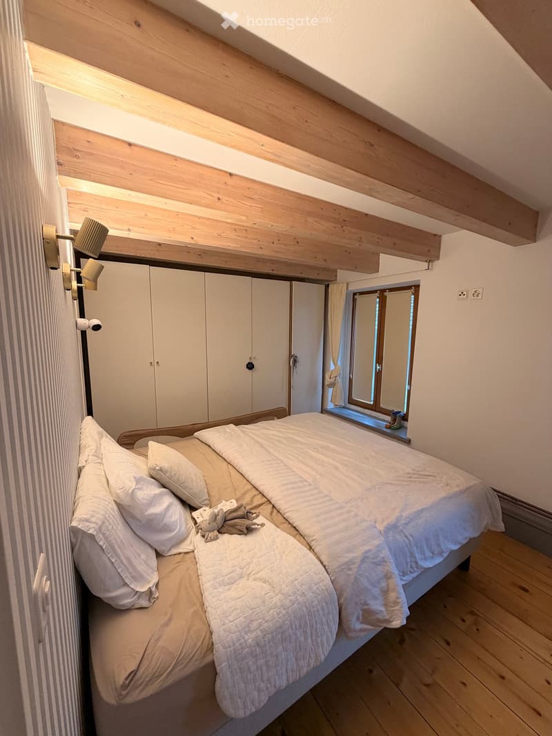 Cozy attic bedroom with wooden slat ceiling, large window, bed with white bedding, and built-in white wardrobe.