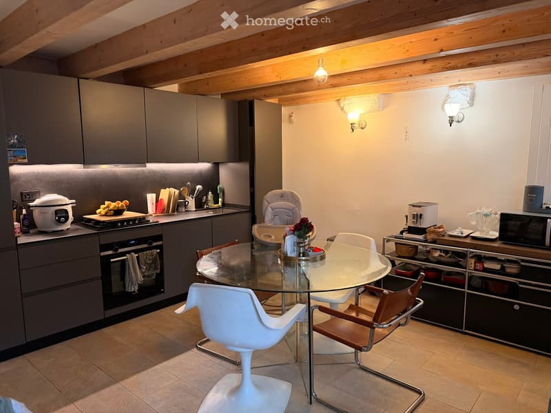 Modern kitchen with grey cabinets, stainless steel appliances, a glass dining table with white and brown chairs, and a baby chair. The kitchen features a wooden ceiling and warm lighting.