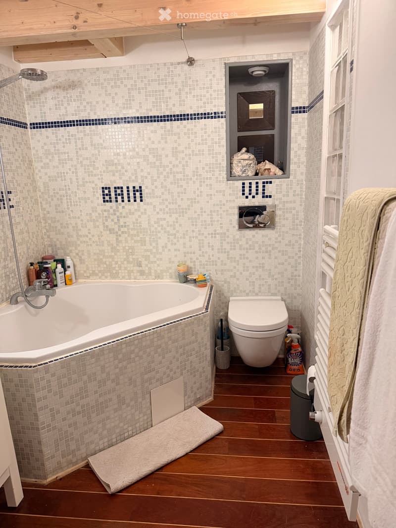 Modern bathroom with a bathtub, toilet, shower, and a built-in shelf holding various items. The walls are tiled with small white tiles and blue accents, and the floor is wooden.