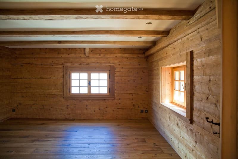 Empty wooden room with exposed beams, wooden walls, and two windows allowing natural light.