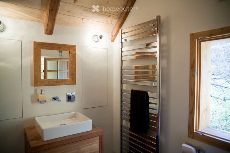 Bathroom with a white sink on a wooden vanity, a framed mirror, a towel rack with a dark towel, and a window showing greenery outside.