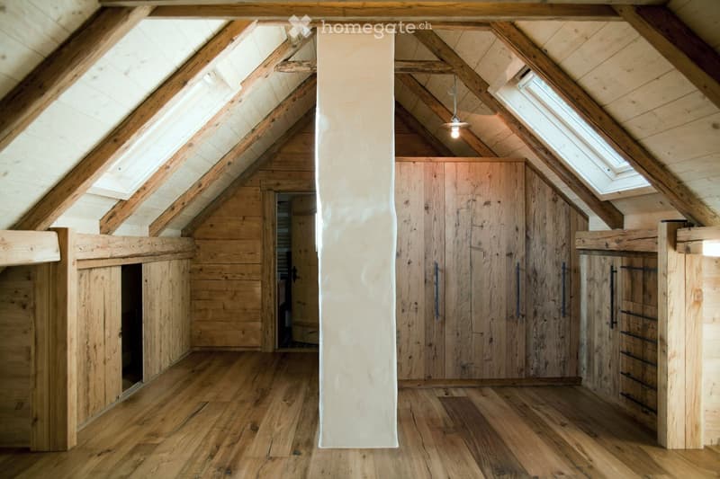 An attic space with wooden beams, exposed wooden walls, and a large wooden wardrobe with multiple doors and drawers. The room features wooden flooring and skylights allowing natural light to enter.