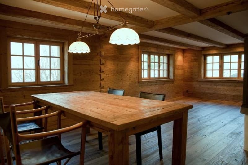 Empty room with wooden walls, exposed beams, large windows, and a wooden dining table with chairs.