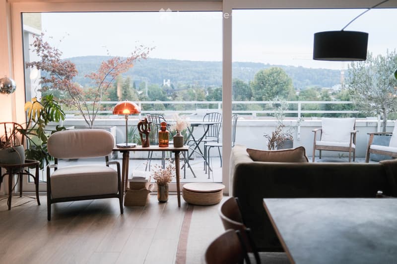 Modern living room with large windows offering a scenic view, featuring a sofa, coffee table, chairs, a lamp, and decorative items.