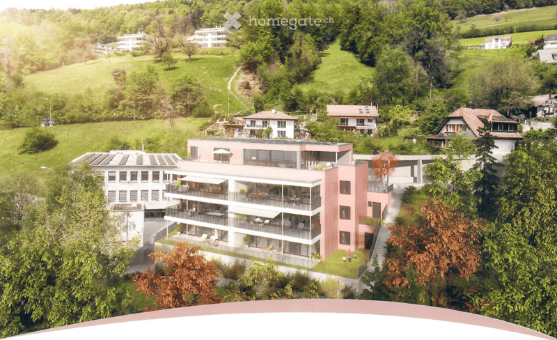 Aerial view of a modern residential complex with multiple buildings, surrounded by lush green hills and trees. The complex includes a prominent pink building with balconies and outdoor seating areas, integrated into a scenic, hilly landscape.