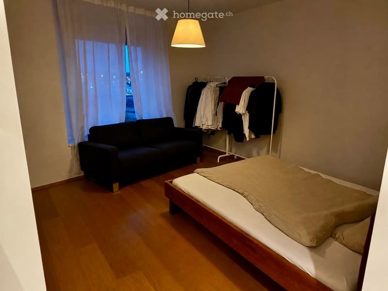 A small, furnished room with a wooden floor, a bed with a beige duvet, a dark-colored sofa, and a clothing rack with hanging clothes.