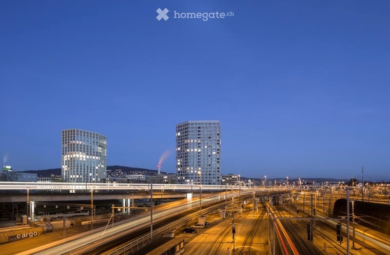 Two modern high-rise buildings with illuminated windows, surrounded by a busy railway network and traffic at dusk.