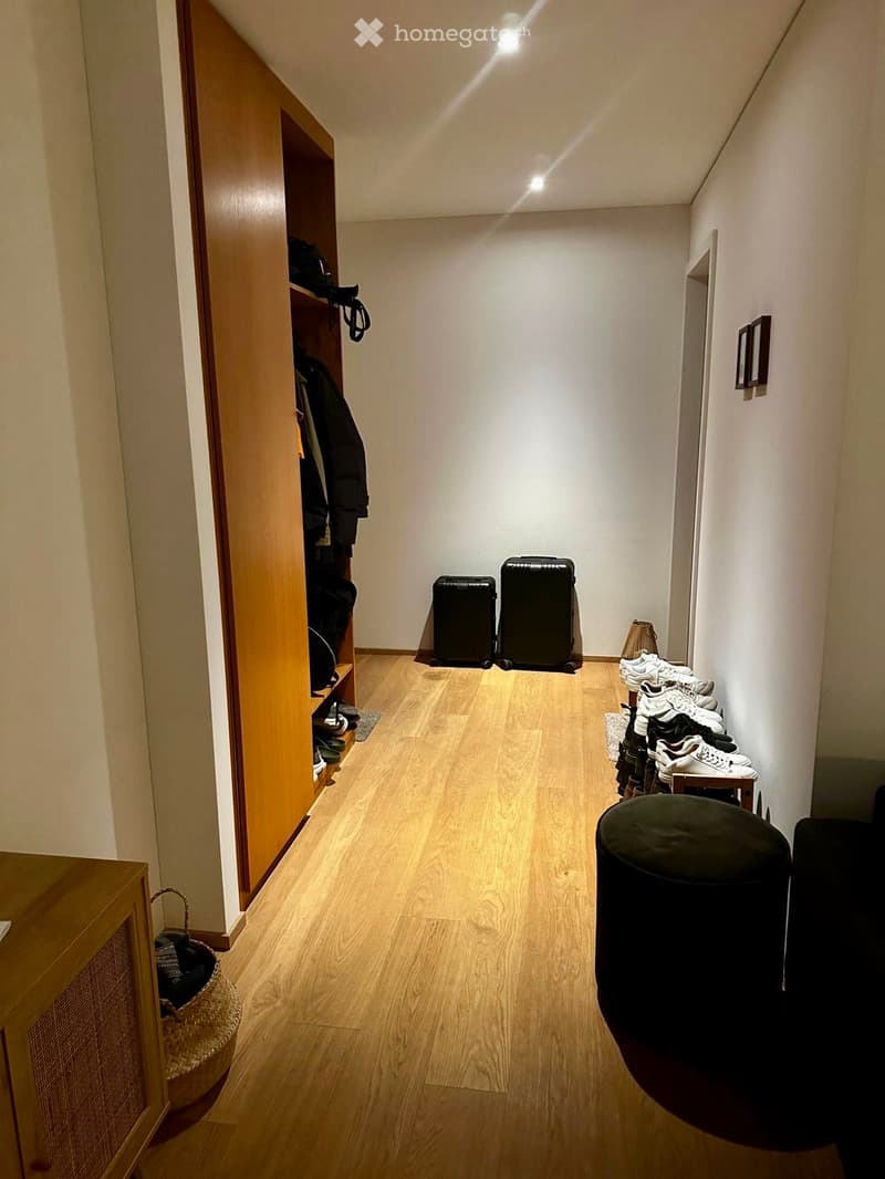 A narrow hallway with wooden flooring, white walls, and a built-in wooden shoe rack on the left side. Two large suitcases are placed in the center, and a stack of shoes is on the right side. A small black stool and a woven basket are also visible.