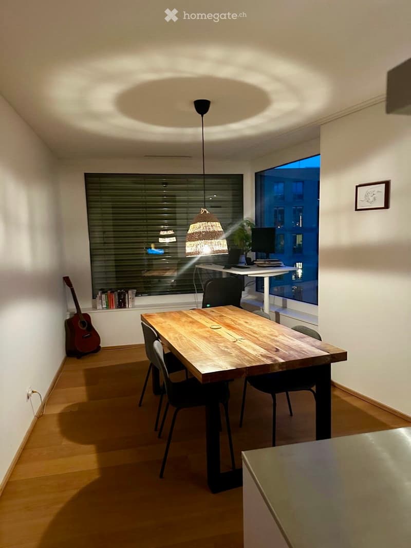 A modern dining area with a wooden table, four chairs, a guitar, a laptop on a desk, and a woven pendant light.