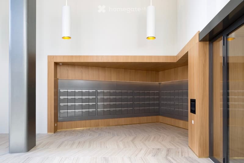 Modern, minimalist hallway with wooden flooring, white walls, and a built-in mail locker system. The space is clean and uncluttered, with pendant lights hanging from the ceiling.