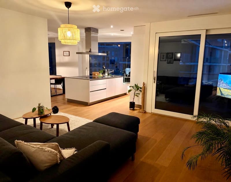 Modern living and dining area with a sectional sofa, ottoman, coffee table, stools, and a kitchen island. Features pendant lighting, a large window, and a TV screen visible through the glass door.