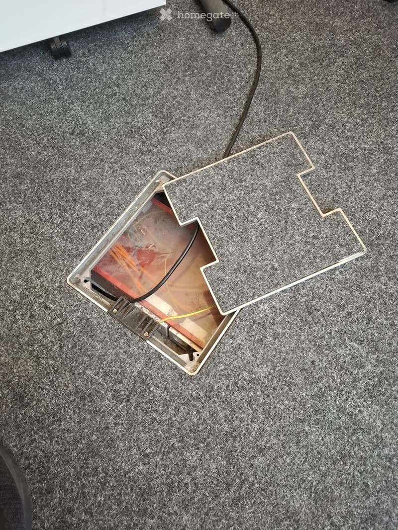 A close-up view of a carpeted floor showing a rectangular hole with exposed wiring and a cable running through it.