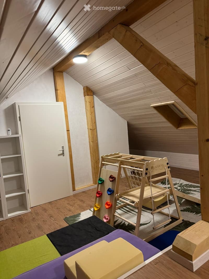 Brightly lit attic room with wooden beams, featuring a rock climbing wall with colorful holds, a play gym structure, foam blocks, a storage cabinet, and a patterned rug.