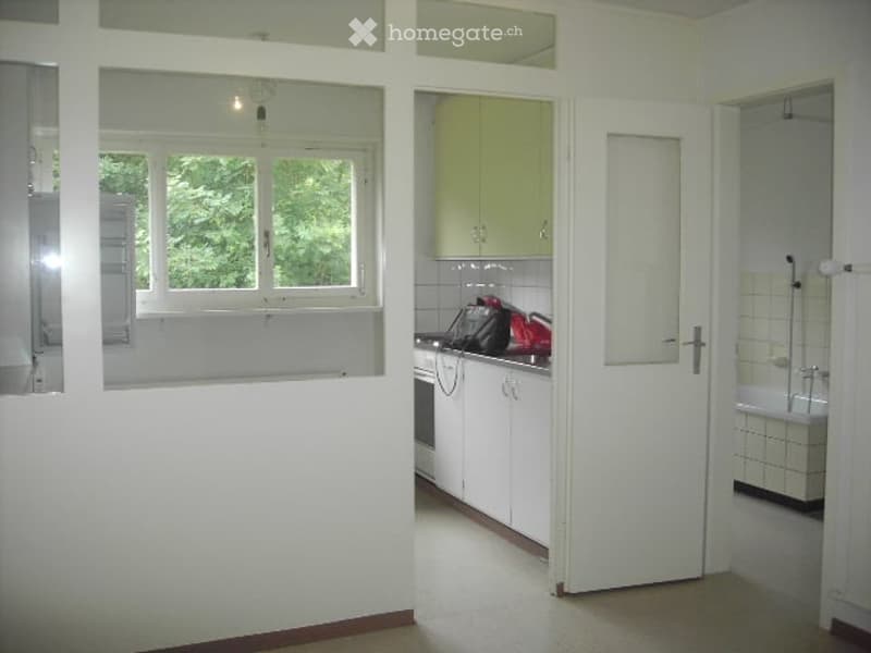 The image shows a modern, minimalist kitchen area with white cabinets, a built-in oven, and a refrigerator. A bathroom is visible through an open door, featuring a white bathtub and tiled walls. The space is well-lit with natural light coming through a la