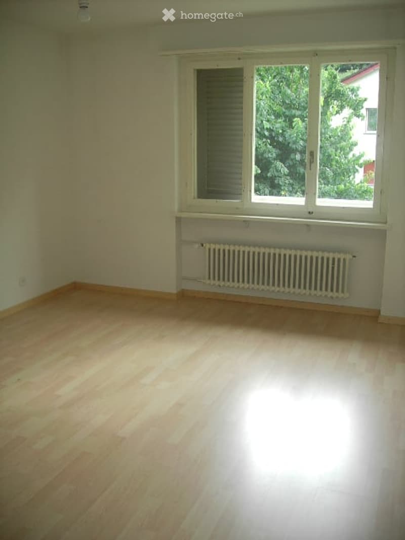 Empty room with wooden flooring, white walls, and a large window with blinds. A radiator is placed below the window.