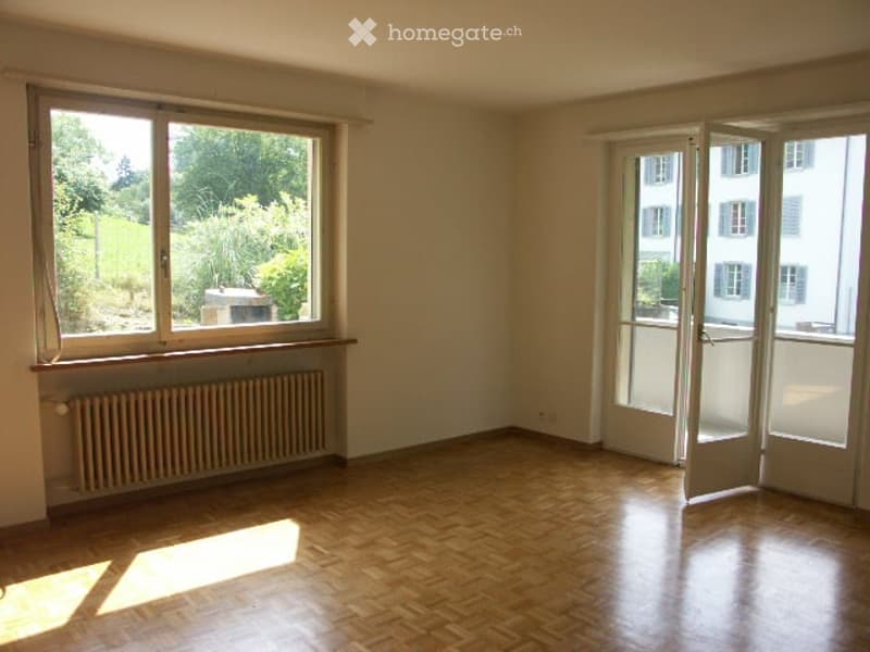 Empty room with wooden flooring, large windows, and a sliding glass door leading to a balcony.