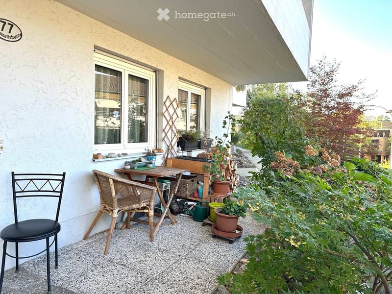 A small outdoor patio area with a wooden table, two chairs, potted plants, and garden tools. The area is surrounded by greenery and has a covered section with a window and a house number sign.