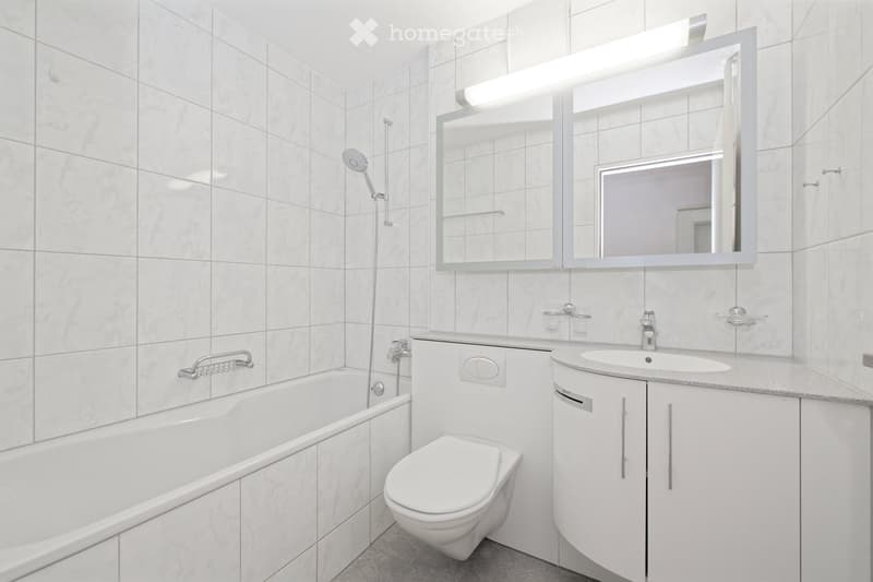 Modern bathroom with white tiles, bathtub, toilet, sink, large mirror with built-in lighting, and modern fixtures.