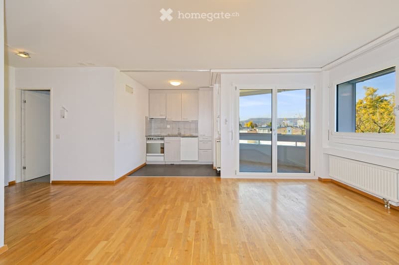 Empty room with wooden flooring, white walls, and large windows. A small kitchen area is visible with white cabinets and appliances.