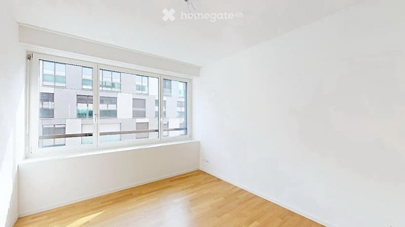 Empty room with white walls, wooden floor, and large window with a view of neighboring buildings.