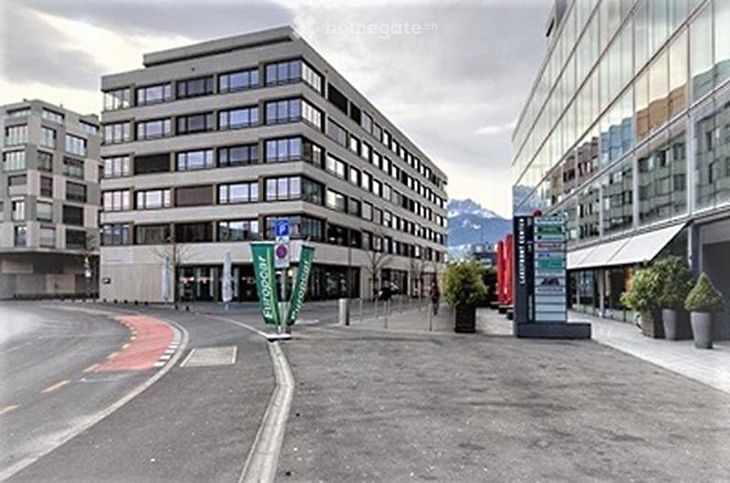 Modern multi-story building with large windows and a contemporary design, situated in an urban area with paved streets and surrounding greenery.