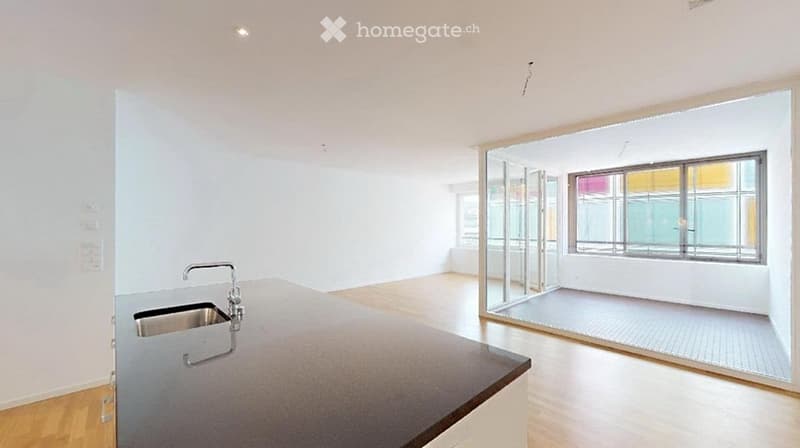 Open-plan unfurnished room with a kitchen counter, sink, and large windows, featuring light wooden flooring and white walls.