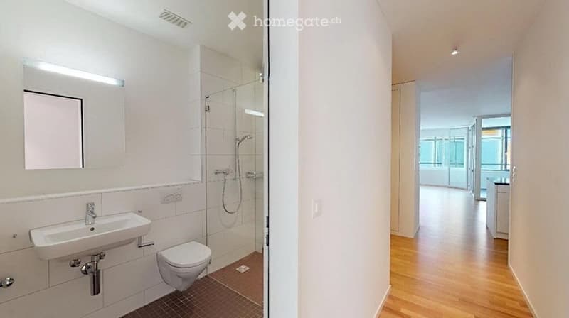 Modern bathroom with white tiles, a sink, toilet, mirror, and a glass-enclosed shower area. The hallway features light wooden flooring and white walls.