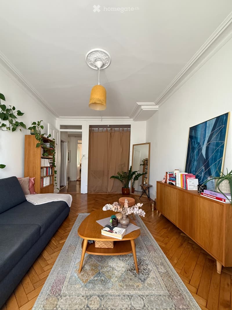 Modern living room with wooden flooring, a blue sofa, a wooden coffee table, a bookshelf, a sideboard, a mirror, and decorative plants.