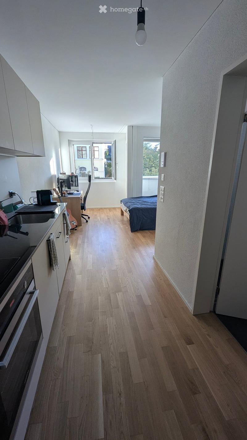 A modern, open-plan living space with a kitchen area, a sleeping area, and a workspace. The kitchen features white cabinets, a black countertop, and stainless steel appliances. The sleeping area includes a bed with blue bedding. The workspace has a desk w