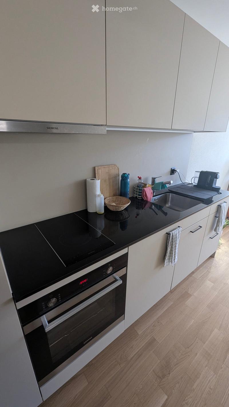 Modern kitchen with white cabinets, black countertops, and wooden flooring. Features an oven, sink, coffee maker, and various small items on the counter.