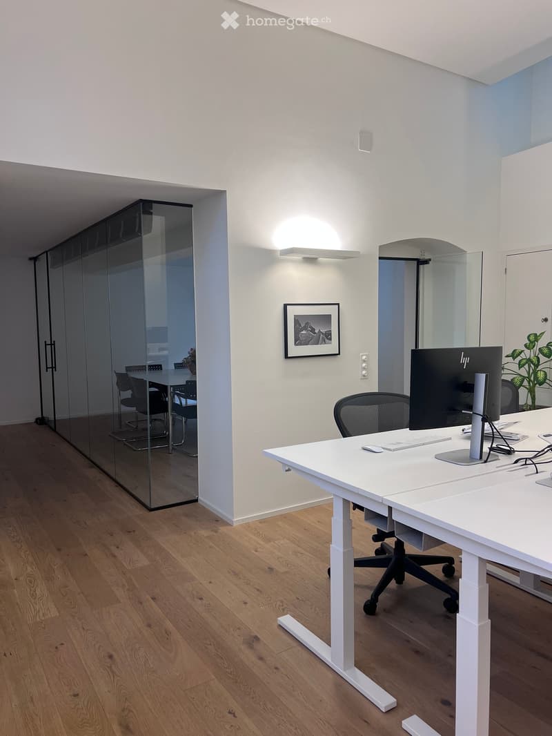 Modern office space with a white standing desk, ergonomic chair, computer monitor, keyboard, mouse, and a framed picture on the wall. A glass partition separates the area from a meeting room with chairs and a table.