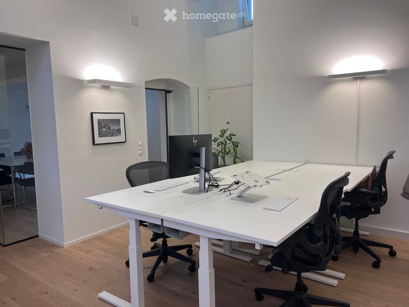 Modern office meeting room with a large white table, multiple chairs, computer setup, and a plant in the corner.