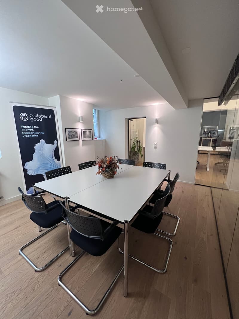 A modern meeting room with a long white table surrounded by black chairs, a flower arrangement in the center, and a large banner on the wall. The room has light wooden flooring and white walls, with a glass partition leading to another room.