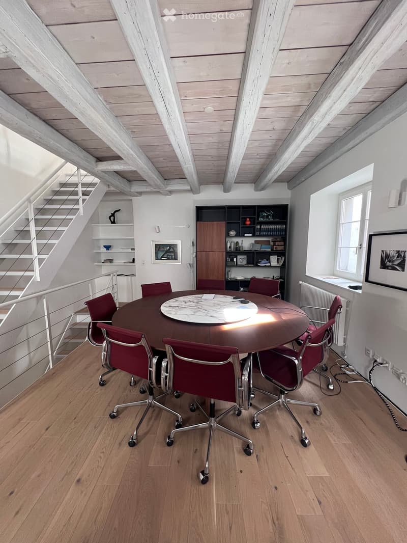 Bright, modern room with exposed wooden beams and slanted ceilings. Features a round wooden table surrounded by red upholstered chairs, a built-in shelving unit, and a staircase leading upwards.