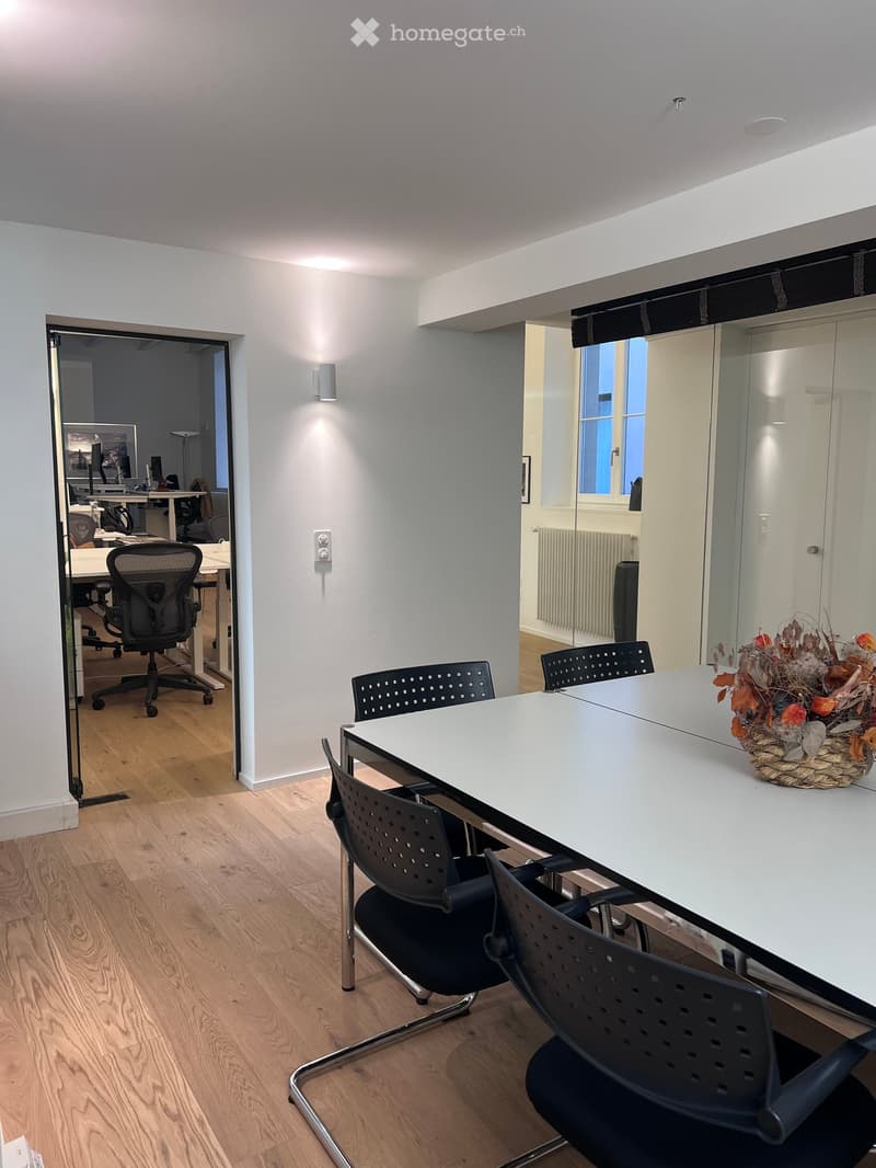 Modern office space with a long white table, black chairs, and a decorative floral arrangement. The room has light wooden flooring and white walls, with a doorway leading to another office area.
