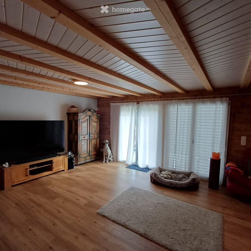 Bright living room with wooden beams, wooden floor, large windows with white curtains, television, dog bed, and a red sofa.